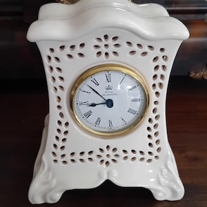 May include: A cream-colored ceramic mantel clock with an intricate openwork lattice pattern. The clock face is white with black Roman numerals, a gold-tone bezel, and the text Royal Creamware printed in the center with a crown icon above it.