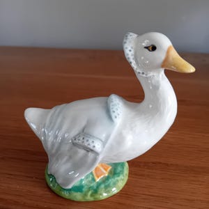 Può includere: Una statuetta di oca in ceramica bianca con un becco giallo e occhi neri. L'oca indossa un vestito a motivi blu e bianchi e sta in piedi su una base verde.