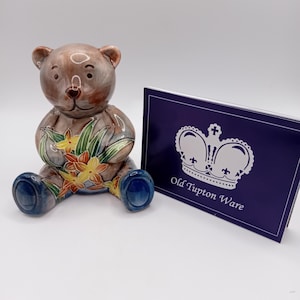 May include: A ceramic teddy bear figurine with a brown body, blue feet, and a floral design on its chest. The bear is seated next to a navy blue card with a crown logo and the text "Old Tupton Ware".