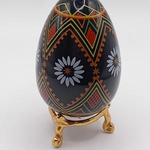 Puede incluir: Un huevo decorativo negro con intrincados patrones en rojo, amarillo y verde, y acentos florales blancos. El huevo se apoya en un soporte dorado con patas curvas, mostrando una mezcla de arte y elegancia.