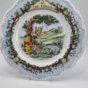 May include: A white ceramic Winnie the Pooh plate with a blue and green border. The plate features a scene from the book "The Rescue" with Winnie the Pooh, Piglet, and Eeyore. The plate is decorated with honey pots around the border.