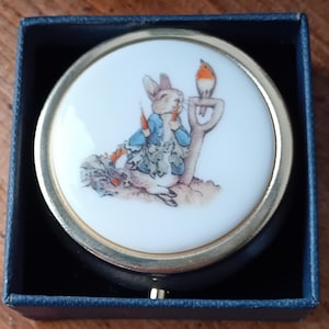 May include: A round, white trinket box with a gold rim, featuring a detailed illustration of Peter Rabbit holding carrots, with a robin perched on a shovel. The box is set in a dark blue presentation box.