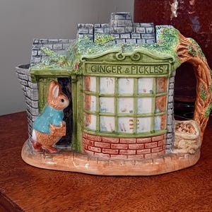 Può includere: Una teiera in ceramica a forma di negozio con un tetto verde e un cartello che recita "Ginger & Pickles". Un coniglio con una camicia blu è in piedi davanti al negozio.