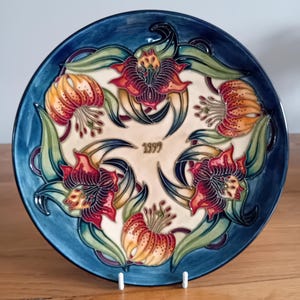 May include: Ceramic plate with a blue border and a floral pattern. The plate has red, orange, and green flowers, with the year "1999" in the centre. The plate is displayed on a white stand.