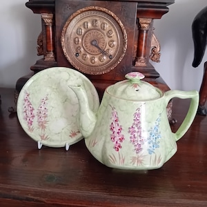 May include: A light green ceramic teapot and matching saucer featuring a painted floral pattern of purple and blue lupine flowers. The teapot has a lid with a small knob, a curved spout, and a handle. The saucer has a matching floral design.