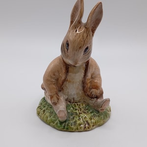 Puede incluir: Una figurita de cerámica marrón y blanca de un conejo sentado sobre una base verde. El conejo tiene orejas largas y mira hacia la izquierda.