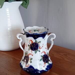 Puede incluir: Un pequeño jarrón de porcelana blanca con ribetes dorados y diseños florales azules y rojos. El jarrón tiene dos asas y un borde festoneado.