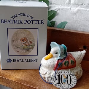 Puede incluir: Una figura de pato de cerámica blanca con un gorro azul y un chal rojo, un pico amarillo y una etiqueta que dice "Peter Rabbit 100". Junto a ella, una caja con "The World of Beatrix Potter" y "Royal Albert".