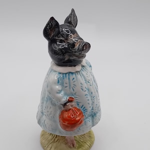 Könnte beinhalten: Eine Keramikfigur eines Schweins mit schwarzem Kopf und Ohren, bekleidet mit einem hellblauen Kleid mit weißem Kragen. Das Schwein hält einen roten Beutel und steht auf einem grünen Sockel.
