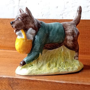 May include: A ceramic figurine of a brown dog wearing a dark green jacket. The dog is depicted in a walking stance, holding a bright yellow basket in its mouth. The base of the figurine is textured and painted in shades of light green and yellow.