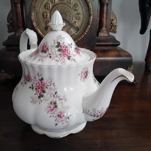 Peut inclure: Une théière en porcelaine blanche avec un corps festonné et un couvercle assorti doté d'un bouton pointu. La théière est ornée d'un motif délicat de roses roses, de feuilles vertes et de fins détails en filigrane gris, avec un liseré doré.