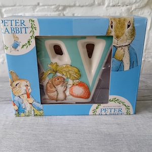Pode incluir: Uma caneca com o tema Peter Rabbit numa caixa azul. A caneca apresenta a letra "V" com uma ilustração de desenho animado de um rato e um morango. A caixa tem imagens de Peter Rabbit e as palavras "Peter Rabbit" impressas.