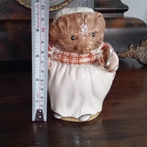 Puede incluir: Una figura de cerámica de un erizo con un delantal blanco y una bufanda a cuadros. La figura marrón y blanca sostiene una pequeña cesta. La figura mide aproximadamente 10 cm de altura.
