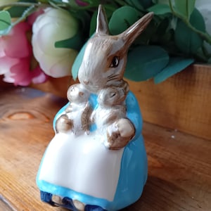 Puede incluir: Una figurita de cerámica de un conejo marrón que lleva un vestido azul y sostiene dos conejos más pequeños. El conejo tiene un delantal blanco y está sentado sobre una superficie de madera.