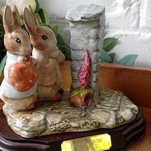 Puede incluir: Figura de cerámica con dos conejos junto a una estructura de piedra. Un conejo lleva un abrigo blanco y una bolsa. La base tiene una placa dorada que dice "Hiding From The Cat". La figura está sobre una base de madera oscura.