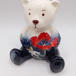 Puede incluir: Figura de oso de peluche de cerámica con cabeza blanca y cuerpo decorado con una amapola roja y un diseño floral azul. Las patas y la base del oso son de color azul oscuro, sobre una superficie blanca.