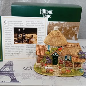 Könnte beinhalten: Miniatur-Häuschen-Figur mit Strohdach, Steinmauern und einer grünen Tür. Das Häuschen ist mit Fässern, Obstkörben und Blumen geschmückt. Teil der Lilliput Lane-Kollektion.