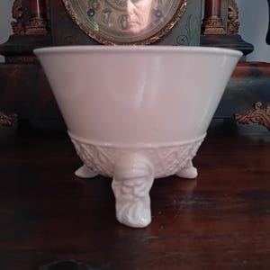 Pode incluir: Uma taça branca de três pernas com uma borda lisa e alargada e uma base texturizada. Cada um dos três pés é esculpido na forma de um rosto barbudo, proporcionando um elemento decorativo distinto à estrutura do recipiente de cerâmica.