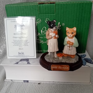 Puede incluir: Figurita de cerámica con un perro y un gato, ambos con delantales. La base lleva la inscripción "GINGER AND PICKLES". Un certificado y una caja verde están en el fondo. La figurita está colocada sobre un libro.