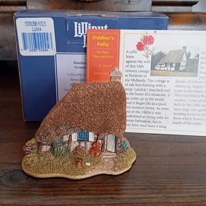 Könnte beinhalten: Miniatur-Figur eines Cottage mit Strohdach, blauen Fensterrahmen und detaillierter Fassade. Die Figur wird von einer Schachtel und einer Karte mit Text über Fiddler's Folly, Pershore, Worcestershire, begleitet. Ein Sammlerstück.
