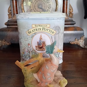 May include: A ceramic figurine of a duck wearing a bonnet and shawl, standing near a wooden fence. The figurine is in front of a tin with "The World of Beatrix Potter" on it. A vintage clock is in the background.