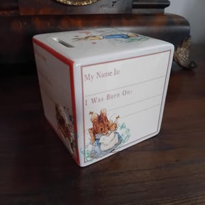 Könnte beinhalten: Eine weiße Keramik-Spardose mit rotem Rand, mit Illustrationen von Peter Hase-Charakteren. Die Box hat einen Schlitz für Münzen und Platz zum Schreiben von "My Name Is:" und "I Was Born On:" an der Seite.