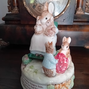Może przedstawiać: Ceramiczna figurka muzyczna przedstawiająca matkę królika trzymającą dwa małe króliczki, z dwoma innymi królikami stojącymi z przodu. Jeden królik ma na sobie niebieską kurtkę i trzyma czerwoną szmatkę w kropki. Tekst na podstawie brzmi: his mother forgave him, to see that he had found his tail and Peter folded up his handkerchief.