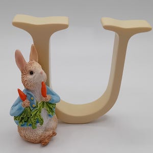 Op de afbeelding: Een decoratief beeldje van een konijn met een blauw jasje en wortels en groen in zijn handen, naast een crèmekleurige letter "U". Het konijnenbeeldje is gedetailleerd met bruine vacht en roze oren. De letter "U" is ongeveer 13 cm hoog.