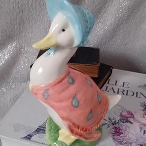 May include: A ceramic duck figurine with a light blue bonnet and a pink shawl adorned with blue teardrop designs. The duck has a yellow beak and is standing on a green base.