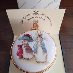 May include: A round white porcelain trinket box with a gold-tone metal rim and hinge. The lid features a colourful illustration of two rabbits, one wearing a green hat and red shawl, the other in a blue jacket. Text reads M.W. Reutter Porcelain Quality since 1948.
