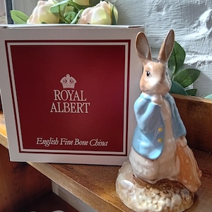 Puede incluir: Una figurita azul y blanca de Peter Rabbit del libro infantil de Beatrix Potter. La figurita está hecha de fina porcelana de hueso y está sentada sobre una base marrón. La figurita sostiene un paraguas amarillo. La figurita está hecha por Royal Albert.