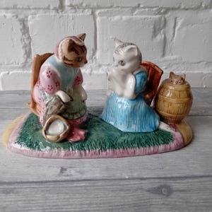 Puede incluir: Una figura de cerámica con dos figuras de gato sentadas en una alfombra, una con un vestido rosa y la otra con un vestido azul. También hay un pequeño barril con un gato asomando. La figura está pintada con detalles y colores.