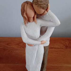 May include: A carved wooden figurine of a couple embracing. The woman wears a white dress, and the man is in a white sweater and brown pants. The figurine has a neutral color palette and a textured finish, suggesting a handcrafted aesthetic.