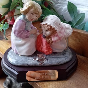 Puede incluir: Figurita de porcelana que representa a una niña y un erizo, ambos vestidos con trajes de época. La figura está montada sobre una base de madera oscura con una placa plateada que dice "Mrs. Tiggy-Winkle and Lucie".