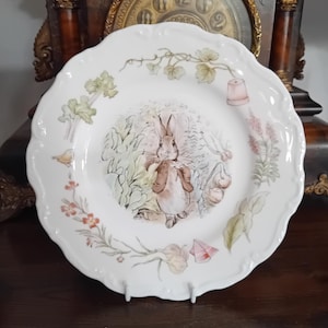 May include: A white ceramic plate with a scalloped edge featuring a central illustration of a brown rabbit wearing a tan jacket. The plate is decorated with botanical motifs including green leaves, small orange flowers, and a terracotta flower pot.