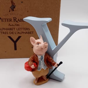以下が含まれることがあります： 茶色のジャケットを着た豚のキャラクターのフィギュアが、赤いバッグと黒い杖を持ち、水色の文字「Y」の隣に立っています。フィギュアはピーターラビットのアルファベット文字セットの一部で、背景に箱が見えます。