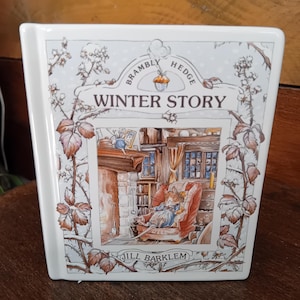 Puede incluir: Una alcancía de cerámica blanca con forma de libro con una ilustración colorida de un ratón en un cómodo sillón junto a una chimenea. El libro se titula "Bramble Hedge Winter Story" de Jill Barklem.