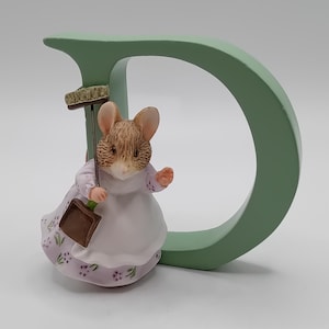 Könnte beinhalten: Figur einer Maus in weißer Schürze und lavendelfarbenem Blumenkleid, die einen Besen und eine Kehrschaufel hält. Die Maus steht neben einem großen, hellgrünen Buchstaben "D". Die Figur hat ein handbemaltes Aussehen.