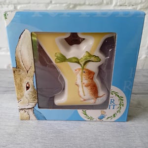 May include: A Peter Rabbit themed gift box featuring a white letter "Y" with a yellow background. A brown and white rabbit holds a green leaf. The box is light blue with Peter Rabbit illustrations.