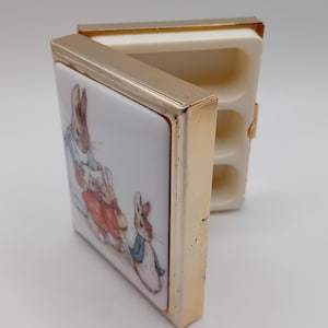May include: A small, rectangular trinket box with a hinged lid. The box features a gold-colored metal frame and a white ceramic surface. The ceramic surface is decorated with a colorful illustration of rabbits in clothing. The interior of the box is off-white.
