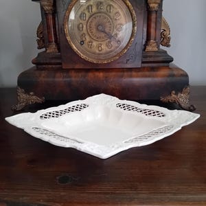 May include: A square white ceramic dish featuring a decorative lattice-work border and embossed floral detailing along the edges. The dish has a smooth, glossy finish and a shallow, concave center, presenting a clean and structured appearance.