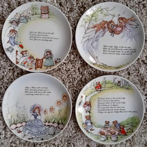 May include: Four decorative porcelain plates with nursery rhyme illustrations. Each plate features a different scene with accompanying text. The plates depict scenes with children, teddy bears, and floral elements, with a soft color palette.
