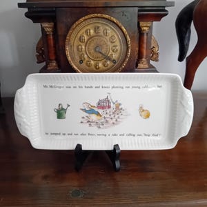 May include: A rectangular white ceramic serving tray with fluted edges and handles. The centre features a Peter Rabbit illustration with a green watering can, a rabbit, and Mr. McGregor, including the text: Mr. McGregor was on his hands and knees planting out young cabbages, but he jumped up and ran after Peter, waving a rake and calling out, 'Stop thief!'