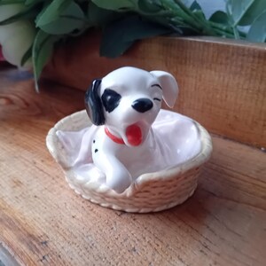 May include: A ceramic figurine of a Dalmatian puppy yawning, with black spots and a red collar, resting in a woven basket. The puppy has one eye closed and is sitting on a white cloth.