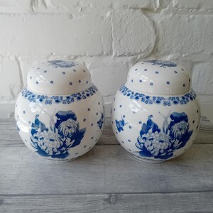 May include: A pair of white ceramic ginger jars with rounded bodies and matching lids. Each jar features a blue floral pattern depicting chrysanthemums and leaves, accented by small blue dots scattered across the surface and a decorative border.