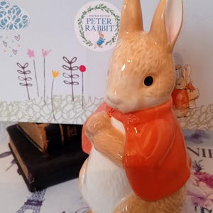 Puede incluir: Una figurita de cerámica de Peter Rabbit con una chaqueta roja. La figurita está de pie sobre una mesa con un libro y un fondo decorativo.