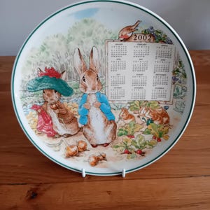May include: A decorative plate featuring illustrations of Peter Rabbit and other characters from the Beatrix Potter stories. The plate has a green border and a calendar for the year 2002. The plate is displayed on a white stand.