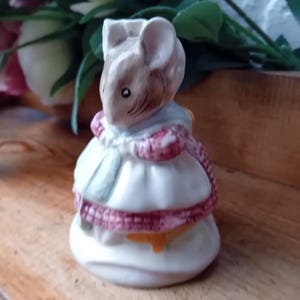 May include: A ceramic figurine of a mouse wearing a white dress with a pink and red checkered trim. The mouse has a light brown face and ears, with a blue collar. The figurine is standing on a white base.