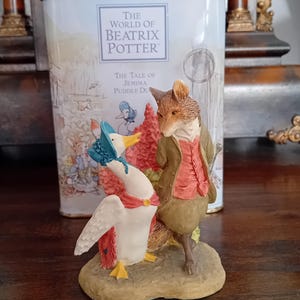Può includere: Statuina in ceramica raffigurante i personaggi di "The Tale of Jemima Puddle-Duck" di Beatrix Potter. La statuina mostra una volpe con un cappotto marrone e un'anatra con un cappello blu e una sciarpa rossa. Una scatola di latta con la scritta "The World of Beatrix Potter" è sullo sfondo.