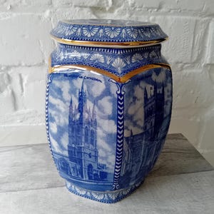 Puede incluir: Un tarro de cerámica con tapa que presenta ilustraciones en azul y blanco de la Catedral de Newcastle y otro edificio. El tarro tiene forma hexagonal con bordes dorados y un patrón repetitivo de conchas en la tapa y el cuello del recipiente.